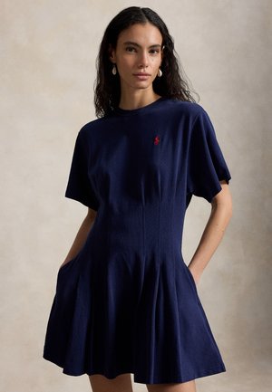 Kvinde med langt krøllet hår iført en mørkeblå kjole med korte ærmer, lommer og et lille rødt logo på brystet, stående foran en ensfarvet baggrund.