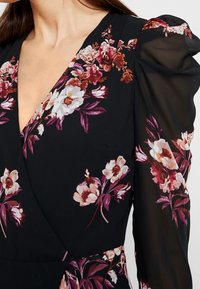 Kvinna iklädd en svart blommig klänning med V-ärmslinje och genomskinliga puffärmar som har rosa och vita blommor.