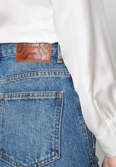 Jeans de mezclilla en azul claro con un acabado texturizado. Presenta una etiqueta de cuero marrón con un diseño intrincado en la cinturilla. Parte de una blusa de tela blanca es visible.