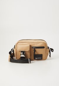 Tan crossbody bag with black accents. Features a zippered compartment, front pocket, adjustable strap, and silver hardware for closure.