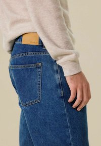 Jeans en denim bleu avec une coupe décontractée, dotés d'une étiquette en cuir beige portant la mention 'SISSY-BOY' et de poches arrière standards.