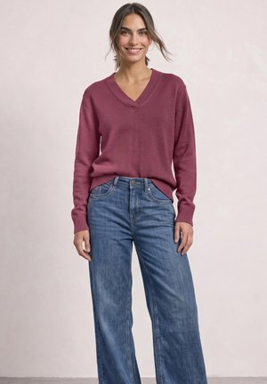 Femme debout portant un pull à manches longues rose foncé à col en V et un jean bleu large sur un fond neutre.