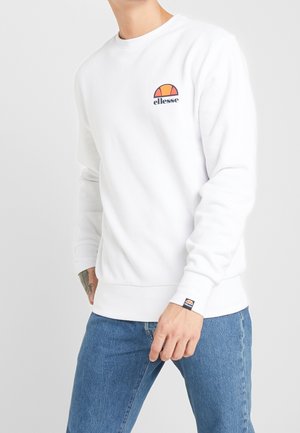 Mand iført hvid ellesse-sweatshirt og blå jeans, med synlig tatovering på venstre håndled og ellesse-logo på brystet og manchet.