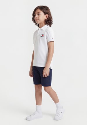 Chlapec s vlasmi po plecia stojaci, oblečený v bielom polo tričku s logom vlajky, námorníckych šortkách, bielych ponožkách a bielych teniskách.