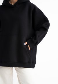 Felpa nera con cappuccio realizzata in tessuto morbido, con tasca frontale e polsini a coste. Indossata sopra pantaloni chiari, vista ravvicinata.