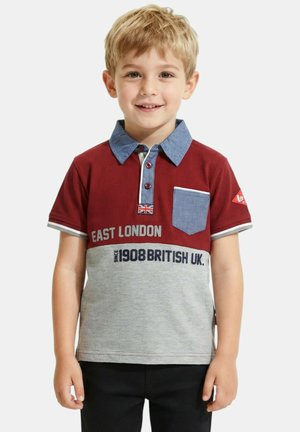 Garçon blond souriant, portant un polo rouge et gris à manches courtes avec un col bleu, une poche poitrine, et le texte "East London 1908 British UK".