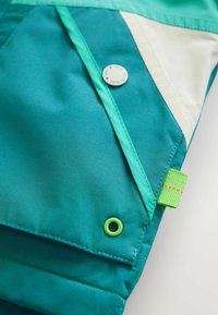 Das Teal-Jackett-Material weist ein kontrastierendes cremeweißes Dreieck, einen grünen Druckknopf und einen dekorativen grünen Riegel mit orangefarbener Naht auf.