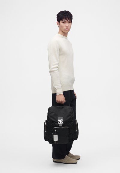 Jeune homme portant un pull blanc et un pantalon foncé tenant un grand sac à dos noir avec plusieurs poches zippées, debout sur un fond uni.