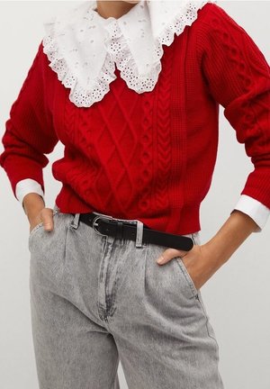 Pull rouge en maille torsadée avec grand col en dentelle blanche porté avec un jean gris taille haute et une ceinture noire, mains dans les poches.