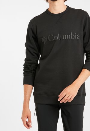 Homme portant un sweat-shirt noir Columbia à manches longues et un pantalon noir, debout devant un fond blanc uni.