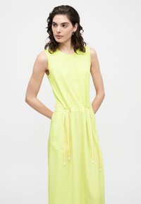 Femme aux cheveux bouclés portant une robe sans manches jaune clair avec une taille à cordon, debout les mains derrière le dos sur fond blanc.