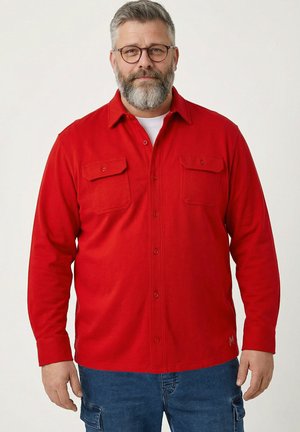 Brodaty mężczyzna w okularach ma na sobie jaskrawoczerwoną koszulę zapinaną na guziki na białym t-shircie i niebieskich dżinsach, stojąc na tle jasnego, jednolitego tła.