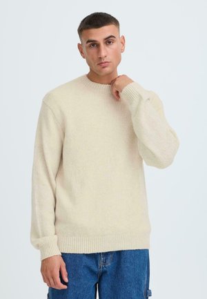 Beige Strickpullover mit Rundhalsausschnitt und gerippten Bündchen. Weiche Textur, am Saum tailliert, getragen mit blauen Jeansshorts.