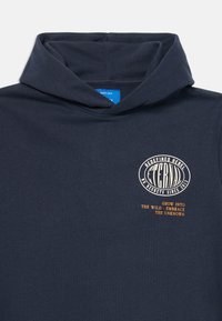 Mørkeblå hættetrøje med en foldet hætte, hvidt oval logo med teksten "Eternal" og "No regrets since 2013." Under dette står der i orange tekst "Grow into the wild."