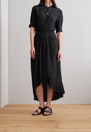 Femme portant une robe midi noire à pois, manches courtes, ourlet asymétrique et sandales noires à brides, debout sur un sol en bois.