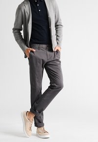 Mężczyzna w szarym żakiecie, granatowym polo, szarych spodniach z mankietami i beżowych sportowych sneakersach, stojący z rękami w kieszeniach na białym tle.