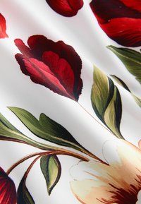 Tissu floral avec des fleurs rouges et crème sur un fond blanc, accompagné de feuilles vertes. Texture lisse et contraste de couleurs vives.