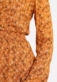 Vaatteiden kietaisu, joka on valmistettu kevyestä, kuvioidusta oranssista kankaasta mustalla kukkakuviolla. Sisältää laskostetun vyötärön ja pitkät, tiukat hihat.