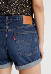 Jeans-Shorts mit umgeschlagenen Säumen, dunkelblauer Farbe, Gesäßtasche mit gesticktem Logo, hellbraunes Lederetikett und kontrastierende Nähtdetails.