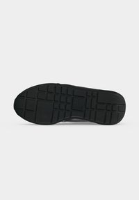 Semelle de chaussure noire avec des motifs de bande de roulement rectangulaires et carrés, montrée de dessous sur un fond clair uni.