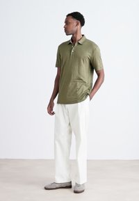 Olivgrön polo med korta ärmar, krage och avslappnad passform, tillsammans med vita vida byxor och grå skor.