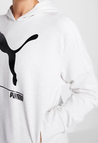 Persona che indossa una felpa bianca Puma con logo nero, mano in tasca, inquadratura a livello del collo e del torso medio, su sfondo semplice.
