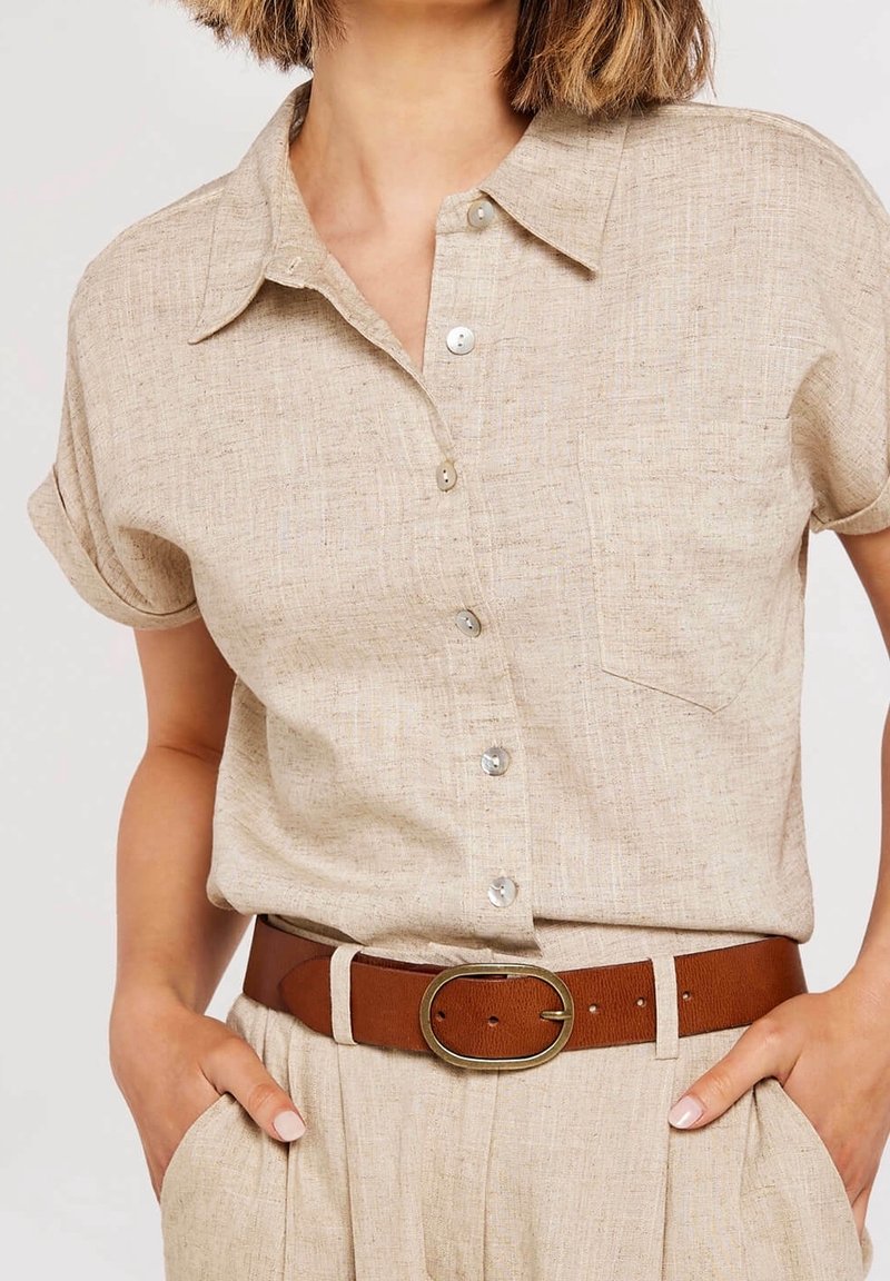 Femme portant une chemise beige à manches courtes avec poche poitrine et pantalon assorti, accessoirisée d'une ceinture marron avec boucle ovale.