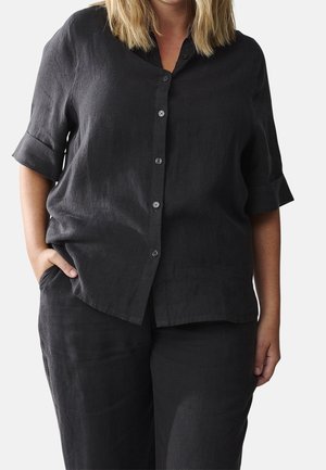 Femme portant une chemise noire à boutons avec manches retroussées et un pantalon noir assorti, une main dans la poche.