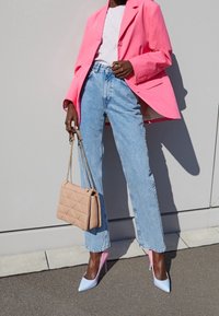 Kvinde iført en klar pink blazer, hvid skjorte, lyseblå jeans, pastelfarvede pink og blå højhælede sko, og holder en beige quiltet håndtaske med kædehanker.