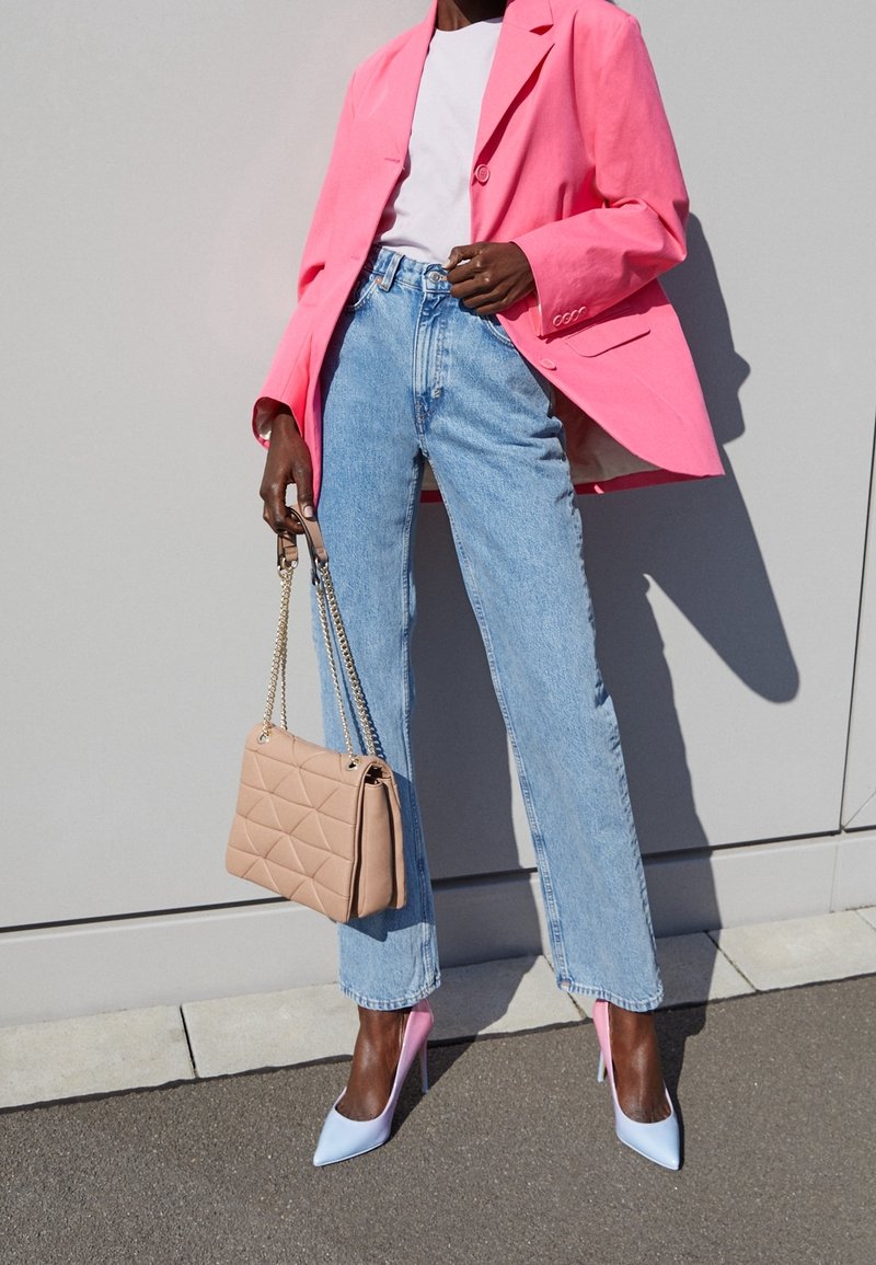 Kvinde iført en klar pink blazer, hvid skjorte, lyseblå jeans, pastelfarvede pink og blå højhælede sko, og holder en beige quiltet håndtaske med kædehanker.
