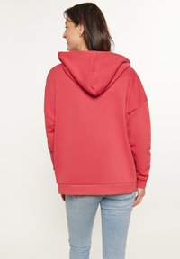 Piros, oversized kapucnis pulóver pamut keverékből készült. Nagy kapucnival, hosszú ujjakkal és bordás mandzsettával rendelkezik, kék farmernadrággal viselve.