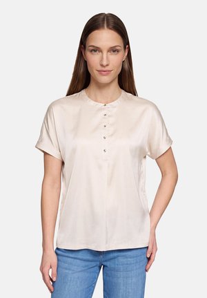 Ung kvinde iført en kortærmet beige satinbluse med knapper foran og blå jeans, stående mod en ensartet lys baggrund.