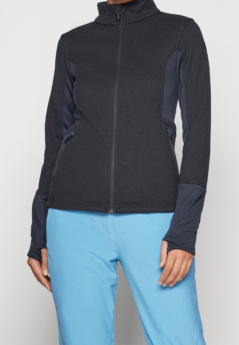 Veste zippée noire texturée avec poignets à trou pour le pouce, portée avec un pantalon bleu clair, montrant la partie avant du haut du corps du cou aux hanches.