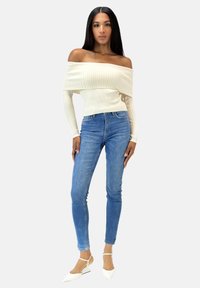 Cremefarbener schulterfreier Ripp-Pullover mit langen Ärmeln, kombiniert mit hoch taillierten blauen Skinny Jeans und weißen spitzen Ballerinas.