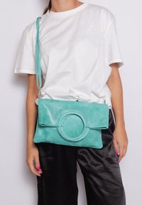 Turquoise leren clutch met een circulaire accent, vlak ontwerp en verstelbare schouderband, gedragen met een wit T-shirt en zwarte satijnen broek.