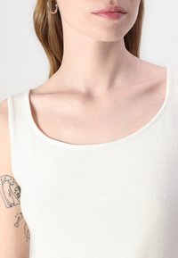 Top in cotone bianco senza maniche con scollatura a girocollo e texture a coste. Presenta il logo in rilievo "GUESS JEANS" sul davanti.
