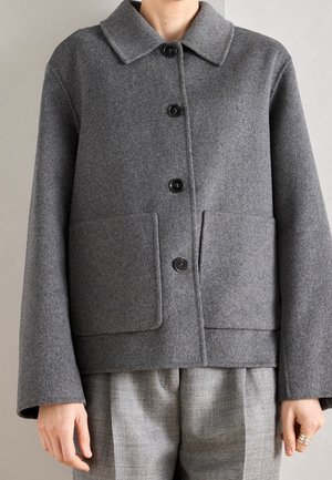 Veste en laine grise avec boutons noirs, col large, poches plaquées à l'avant, portée sur un pantalon à carreaux gris, montrant le torse et les bras.