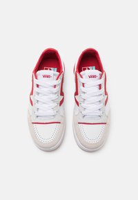 Pár bílých sneakers Vans s červenými akcenty, bílými tkaničkami a perforovanou špičkou, zobrazené shora na jednoduchém bílém pozadí.