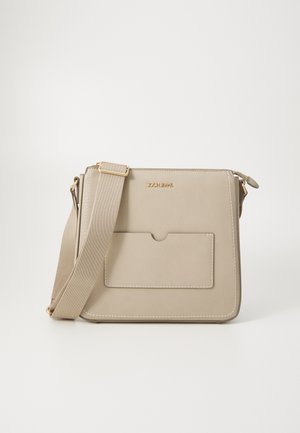 Sac bandoulière beige en matériau lisse, avec une grande poche avant, des accessoires en or et des détails de couture blanche contrastante.