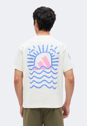 Mężczyzna noszący białą koszulkę z niebieską falą i promieniami słonecznymi oraz różowym logo Adidas na plecach, w połączeniu z oliwkowozielonymi spodniami.