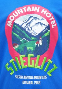 Modrá bavlnená tričko s farebnou grafikou hôr a hotela. Text obsahuje "Mountain Hotel" a "Stieglitz," s vibrantnými ružovými a zelenými akcentmi.