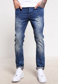 Vaqueros de mezclilla azul con un acabado desgastado, estilo de pierna recta, diseño de cinco bolsillos y dobladillos enrollados, combinados con zapatillas blancas.