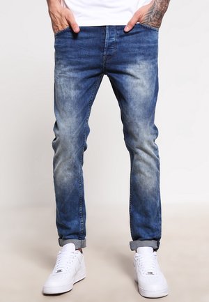 Vaqueros de mezclilla azul con un acabado desgastado, estilo de pierna recta, diseño de cinco bolsillos y dobladillos enrollados, combinados con zapatillas blancas.