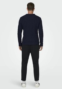Marineblauwe gebreide trui met een kabelpatroon, ronde hals en ribgebreide manchetten. Draagt het met zwarte broek en witte sneakers, waarbij de achterkant zichtbaar is.