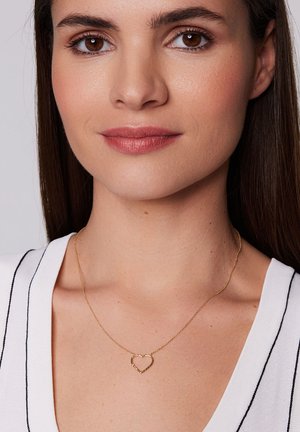 Złoty naszyjnik w kształcie serca na delikatnym łańcuszku, z prostym wzorem i polerowanym wykończeniem, noszony na białej bluzce w czarne paski.