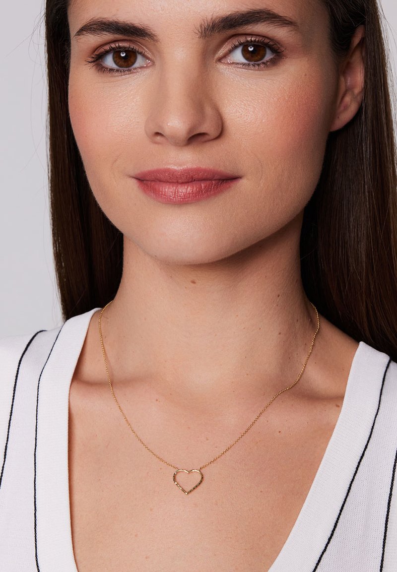 Złoty naszyjnik w kształcie serca na delikatnym łańcuszku, z prostym wzorem i polerowanym wykończeniem, noszony na białej bluzce w czarne paski.