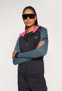 Leichte, grau-gefärbte Jacke mit pinker Kapuze und Akzenten. Sie verfügt über lange Ärmel und eine mit Reißverschluss verschließbare Brusttasche für Stauraum.