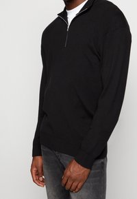 Pull noir à fermeture quart de zip en tissu tricoté texturé, doté d'un col montant, de manches longues et d'un ourlet ajusté. Porté sur une chemise blanche.
