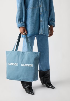 Sac fourre-tout en tissu bleu clair avec le texte blanc "SAM Sø" des deux côtés. Doté de doubles poignées et d'une forme rectangulaire simple.