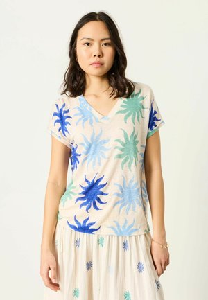 Jonge vrouw die een beige T-shirt met V-hals en een rok draagt, beide met blauwe en groene zonnestraalpatronen, staand tegen een eenvoudige witte achtergrond.